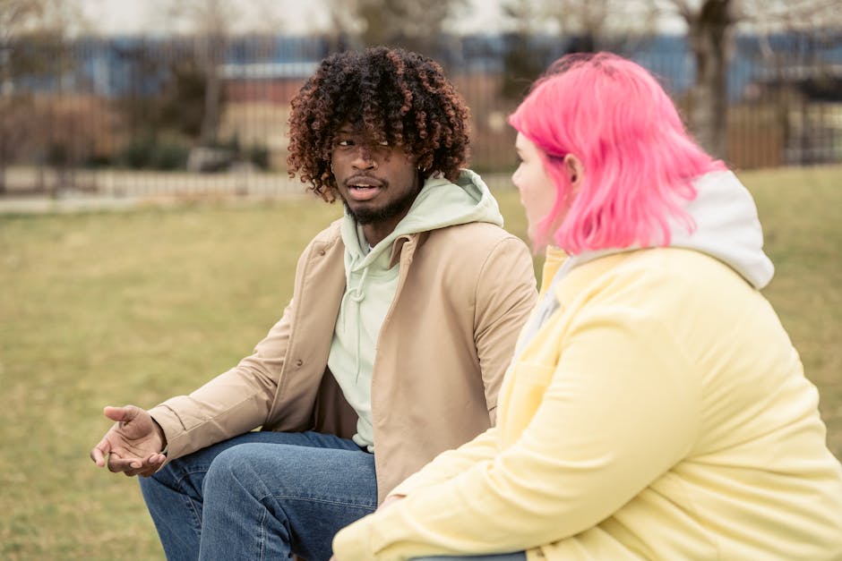 Diverse couple in outerwear looking at each other and having conversation while spending time in park with grassy lawn on street on blurred background